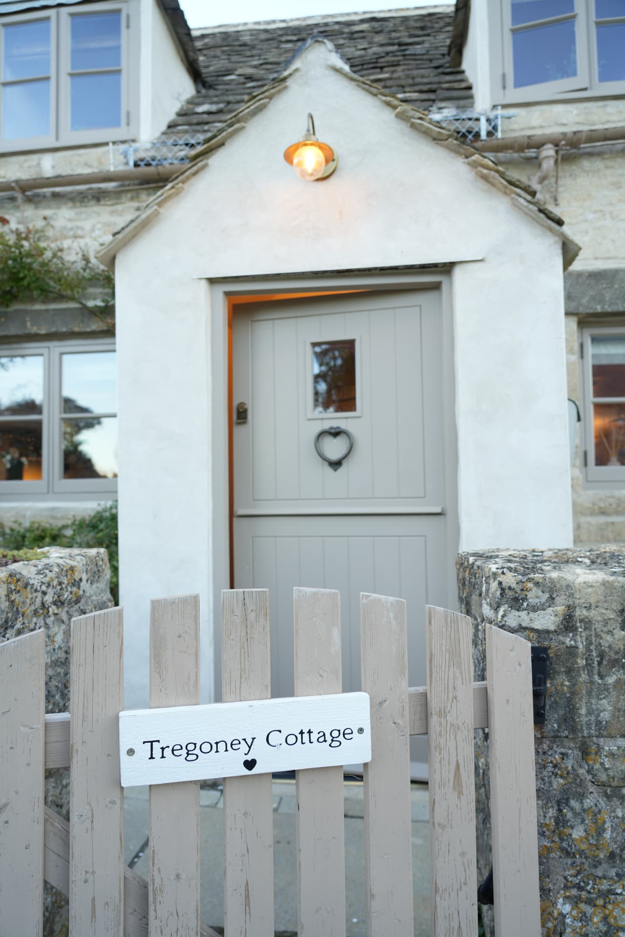 The gate and lit porch of a honey-stone cottage at dusk
