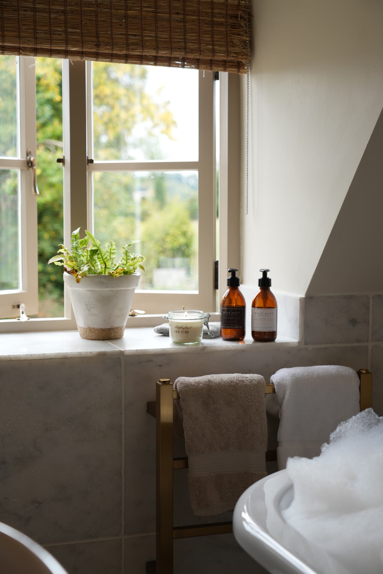 Marble window sill with a fern, lit candle and amber bath bottles above a bubble bath